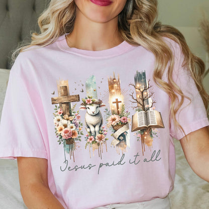A woman wearing a pink t-shirt with a graphic design featuring a cross, a lamb, and a book, along with the text &quot;Jesus paid it all&quot;.