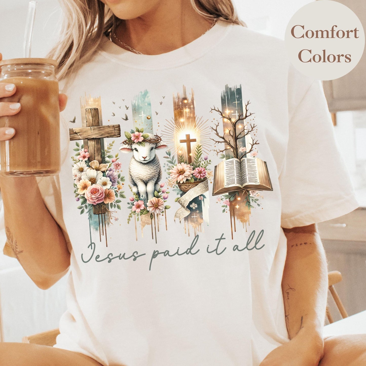 A person wearing a white t-shirt with a graphic design featuring a cross, flowers, and a book, along with the text &quot;Jesus paid it all&quot;.