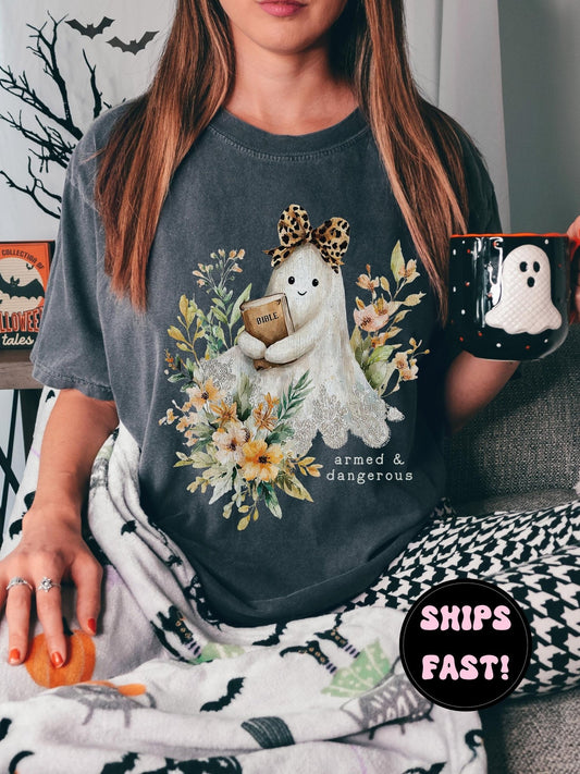 A woman wearing a t-shirt with a graphic of a ghost holding a book and surrounded by flowers, sitting on a couch and holding a mug.