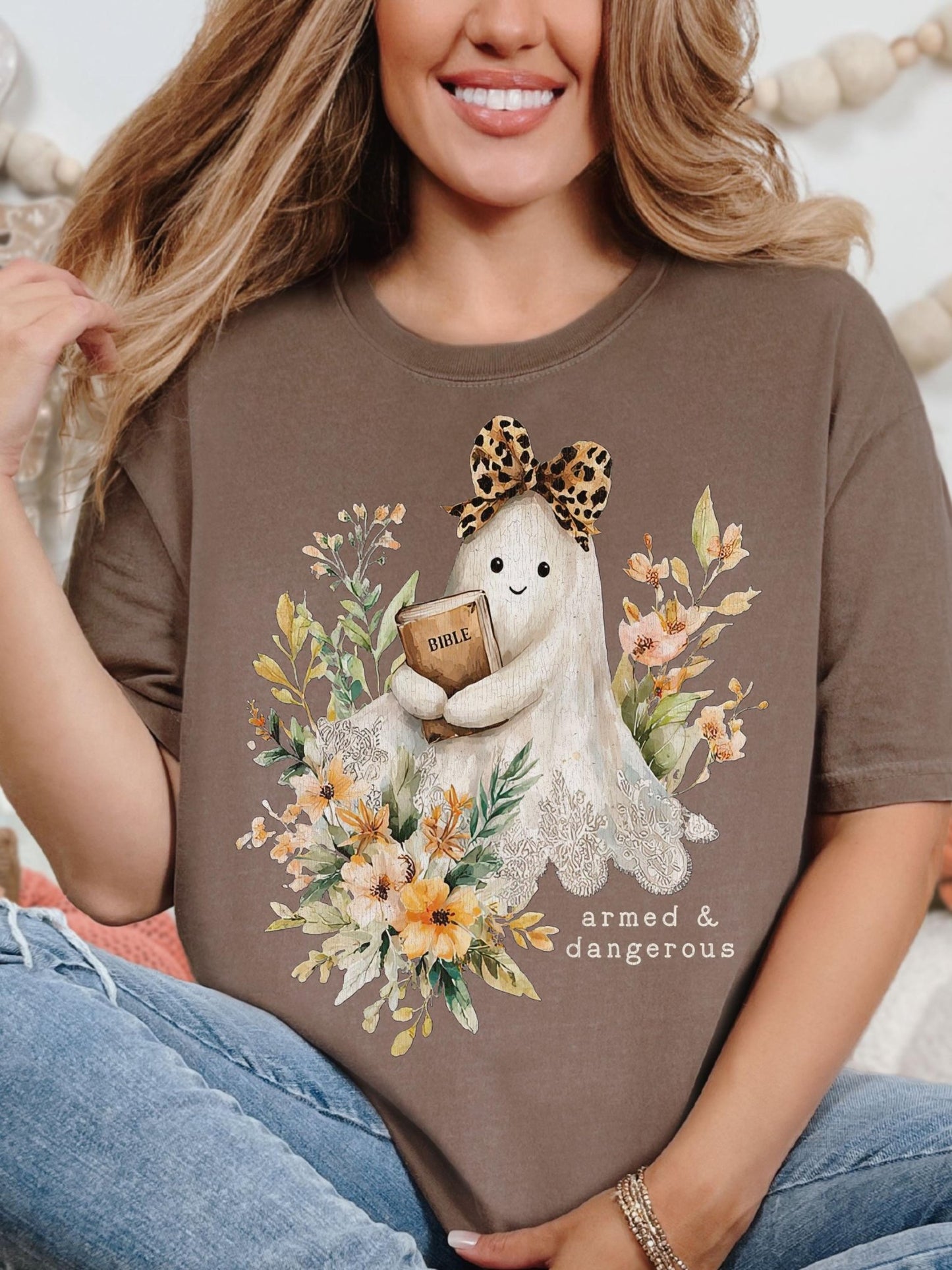 A woman wearing a brown t-shirt with a graphic design of a ghost holding a Bible and surrounded by flowers.