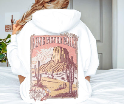 Love Never Fails pink Christian hoodie with desert sunset graphic