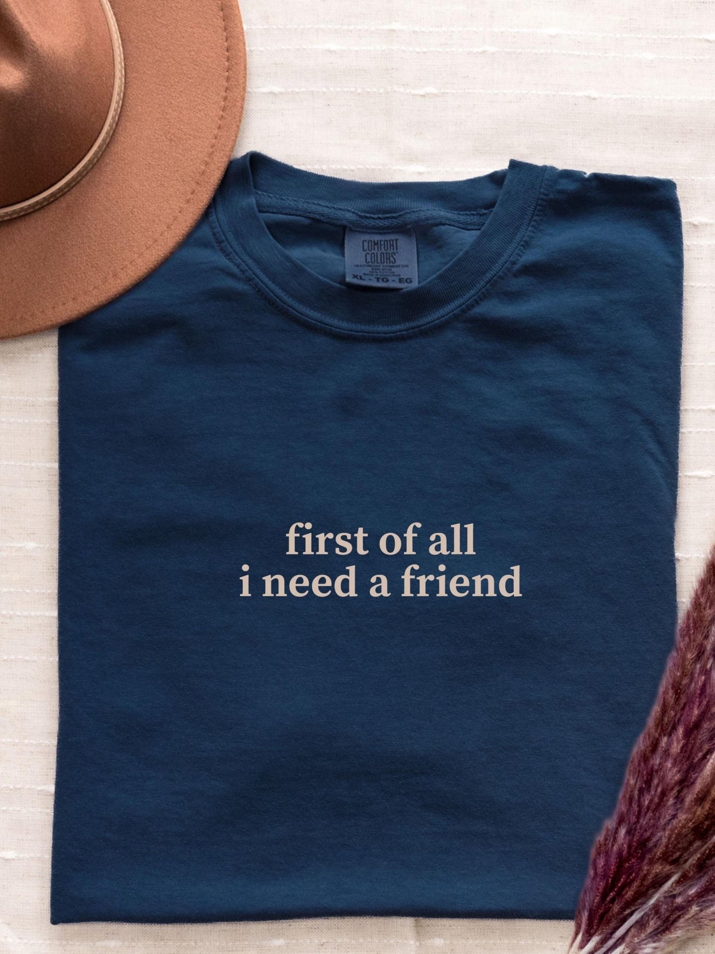 A navy blue t-shirt with the text &quot;first of all I need a friend&quot; printed on it, along with a brown hat and some pink flowers.