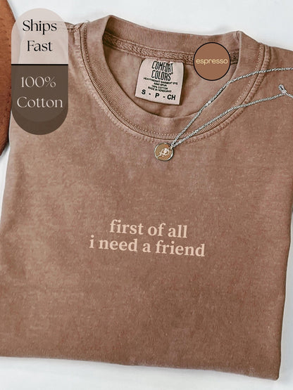 A brown t-shirt with the text &quot;first of all I need a friend&quot; printed on it, along with a small charm necklace.