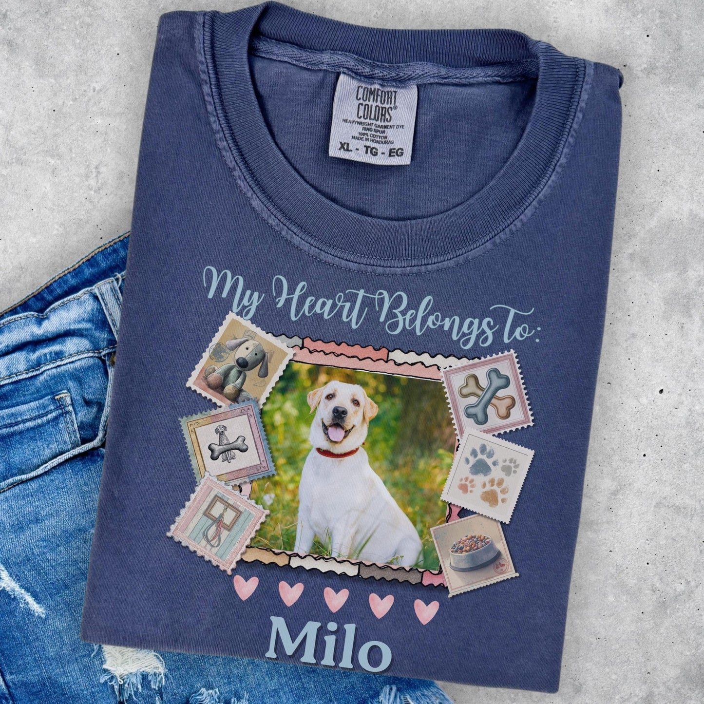 A blue t-shirt with a printed design featuring a white dog surrounded by various dog-related items, such as bones and paw prints, along with the text &quot;My Heart Belongs to Milo&quot;.