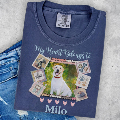 A blue t-shirt with a printed design featuring a white dog surrounded by various dog-related items, such as bones and paw prints, along with the text &quot;My Heart Belongs to Milo&quot;.