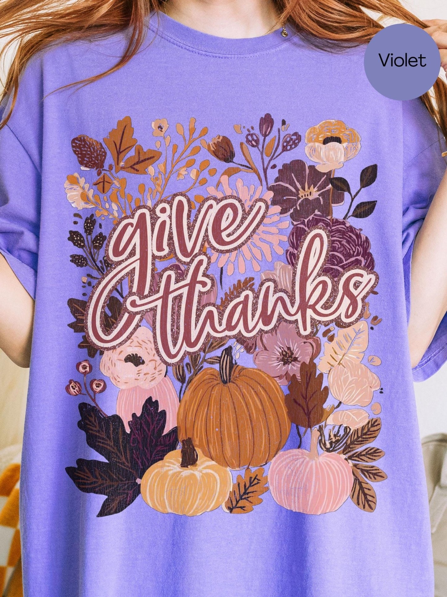A person wearing a purple t-shirt with a graphic design featuring autumn flowers, pumpkins, and the text &quot;Give Thanks&quot;