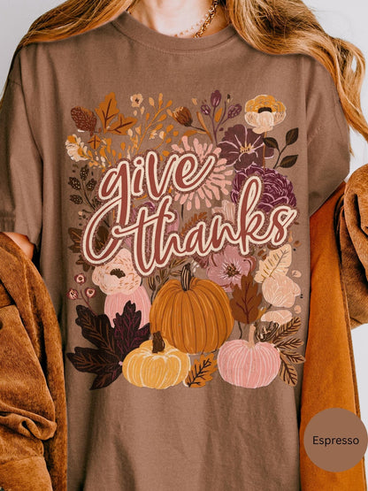 A t-shirt with a graphic design featuring a floral pattern and various autumn-themed elements, including pumpkins, leaves, and flowers. The text &quot;give thanks&quot; is prominently displayed in the center of the design.