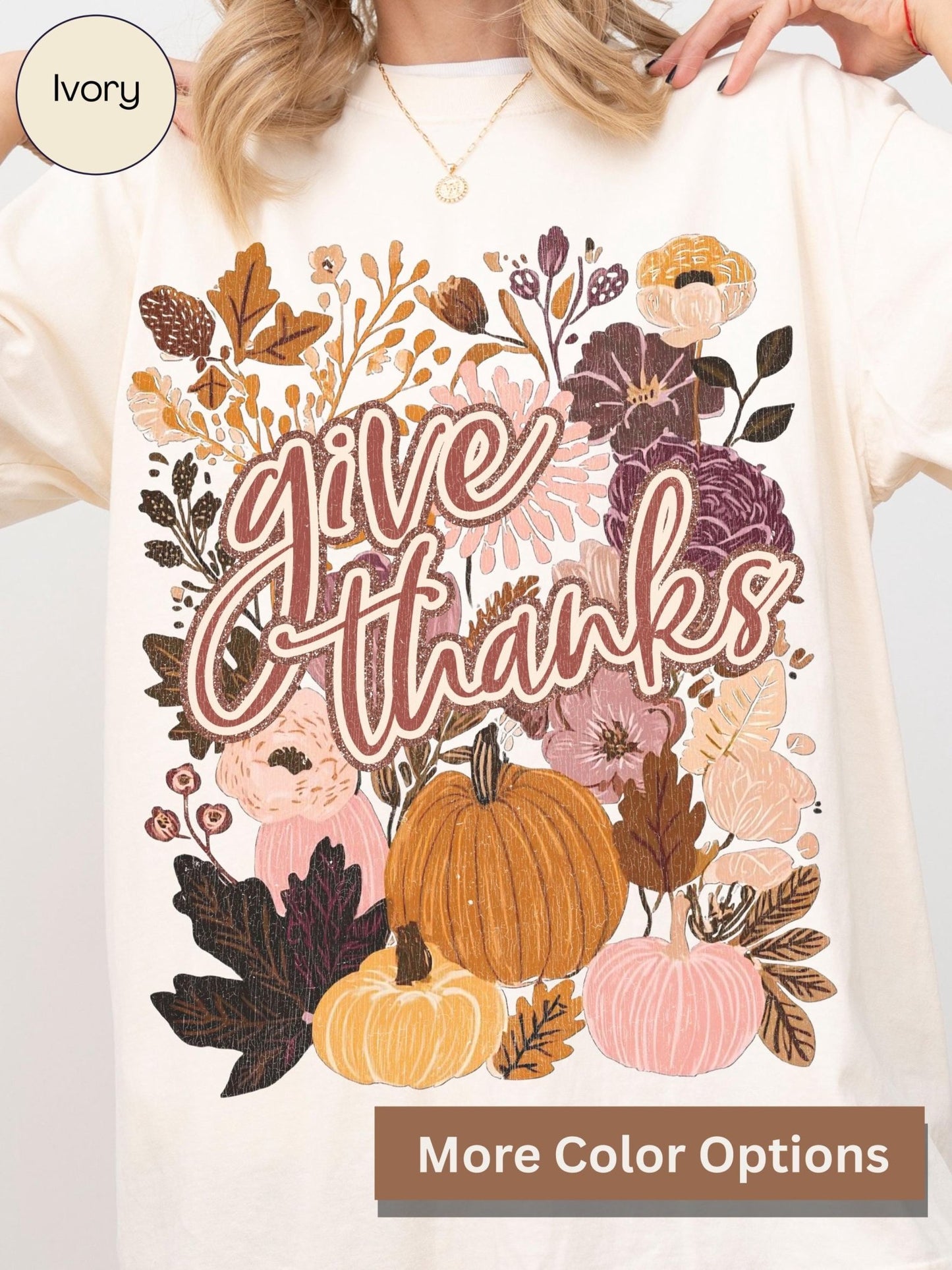 A white t-shirt with a colorful graphic design featuring autumn leaves, pumpkins, and flowers along with the text &quot;Give Thanks&quot;.