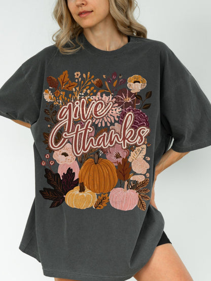 A woman wearing a gray t-shirt with a graphic design featuring autumn flowers, pumpkins, and the text &quot;Give Thanks&quot; printed on it.