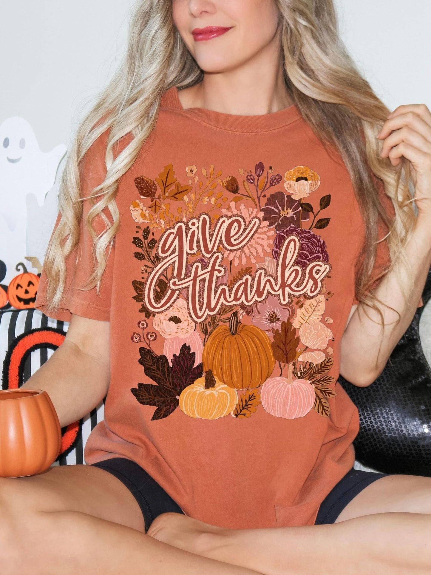 A woman wearing a t-shirt with a floral design and the text &quot;Give Thanks&quot; is sitting on a couch.
