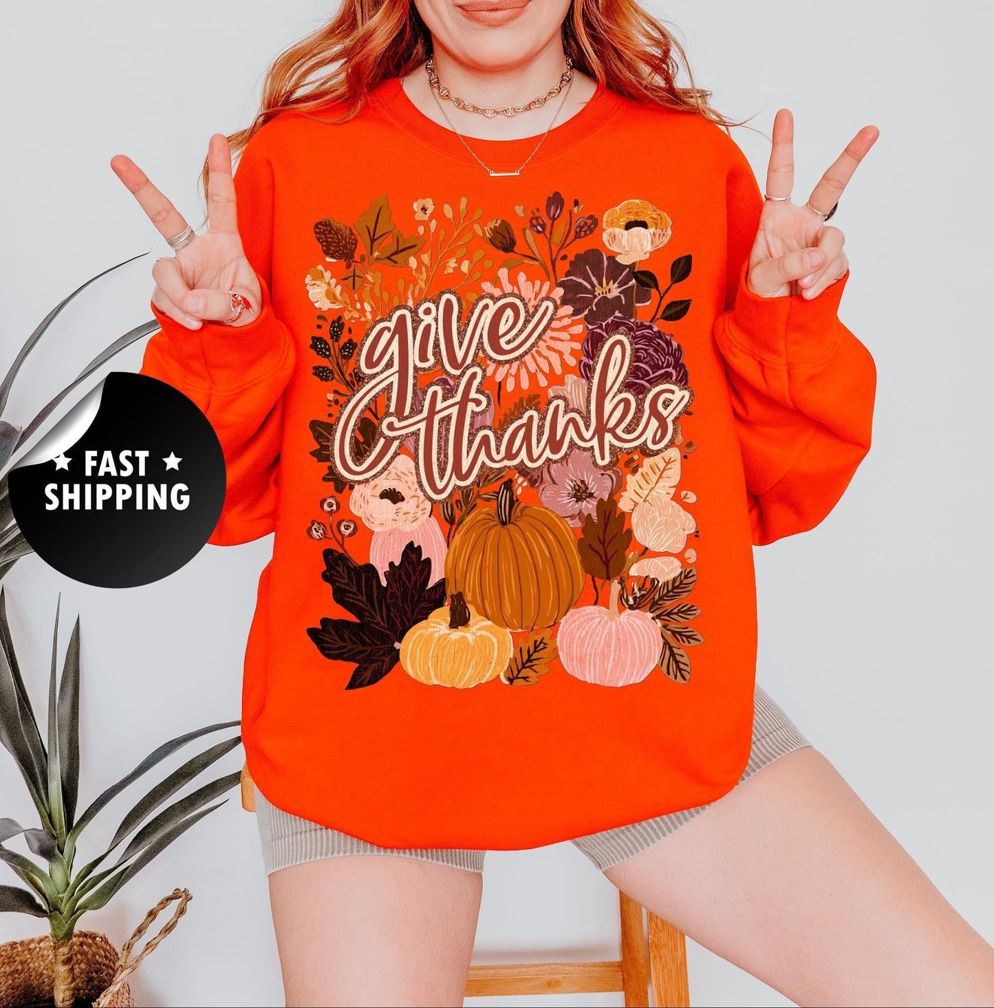 A woman wearing an orange sweatshirt with a floral design and the text &quot;Give Thanks&quot; is making a peace sign with her fingers.