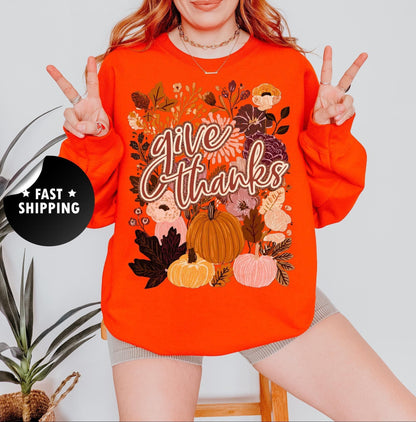 A woman wearing an orange sweatshirt with a floral design and the text &quot;Give Thanks&quot; is making a peace sign with her fingers.