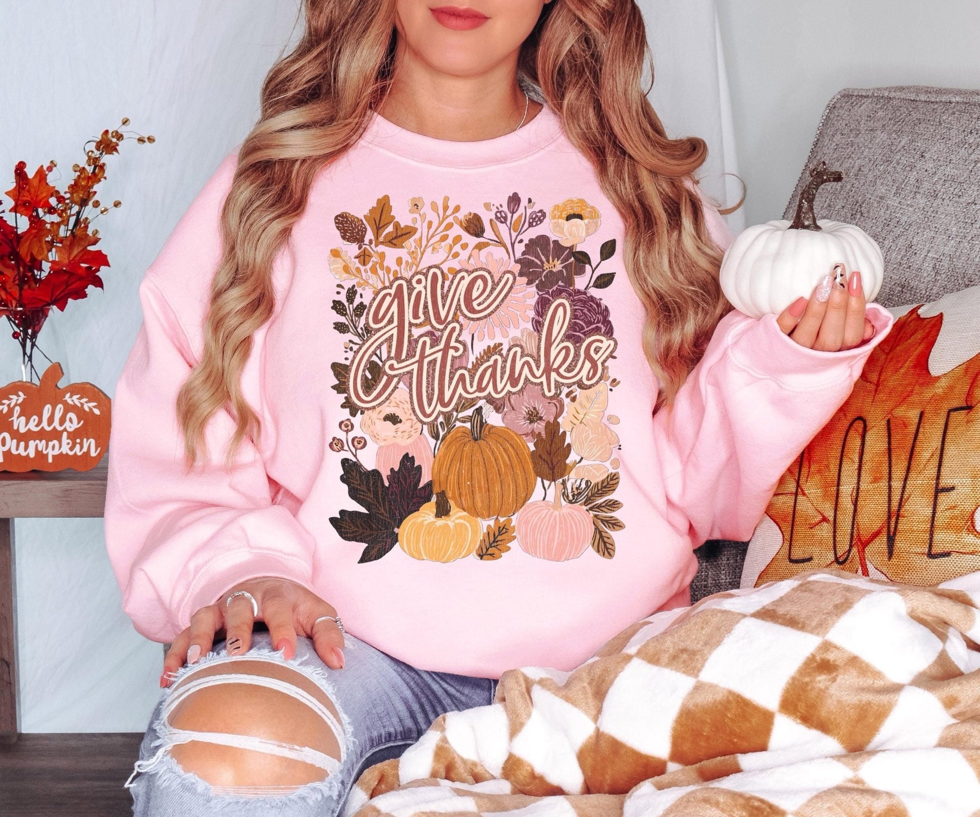 A woman wearing a pink sweatshirt with a pumpkin and fall design, sitting on a couch with a pumpkin pillow and a checkered blanket.