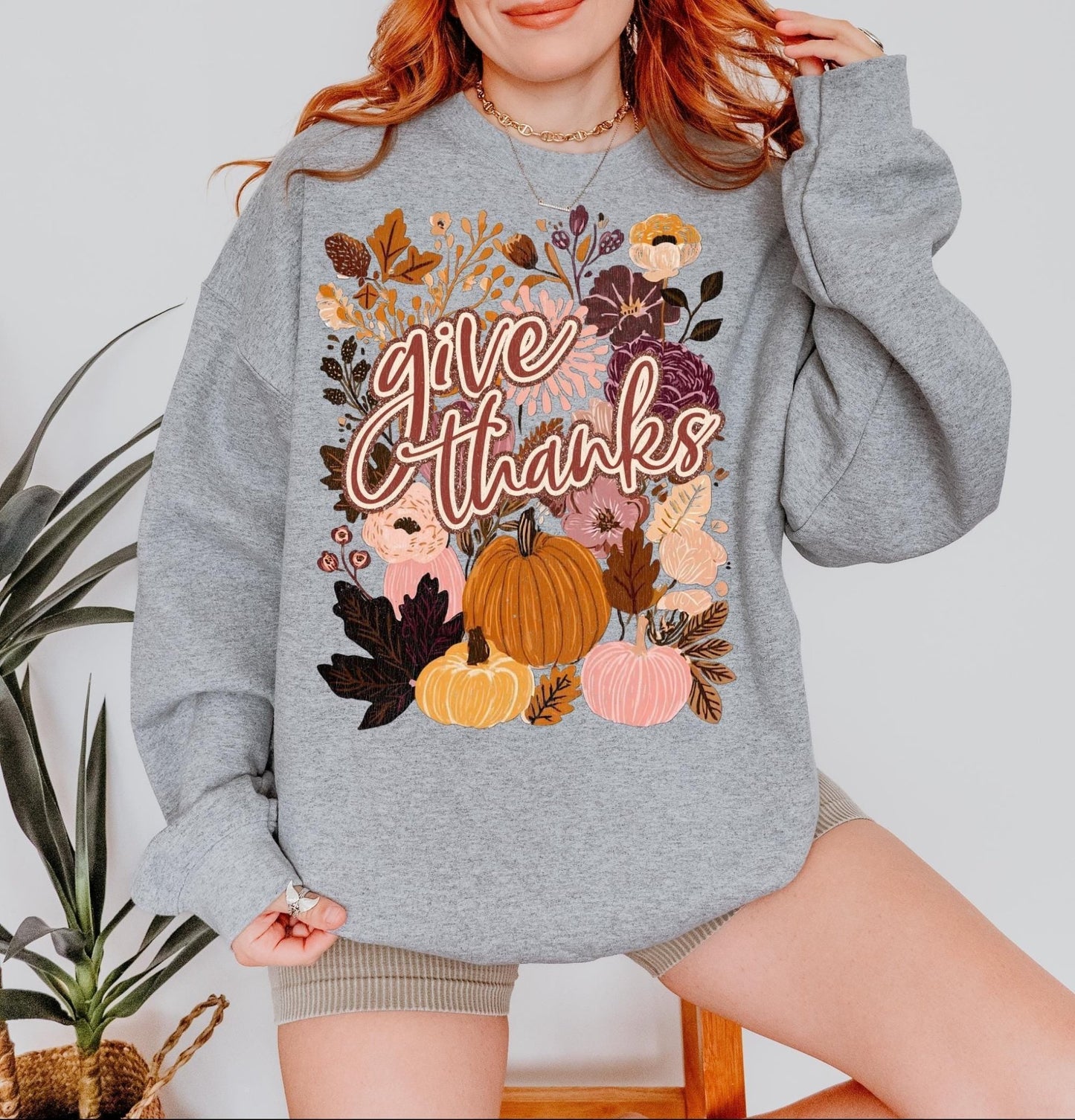 A woman wearing a grey sweatshirt with a graphic design of autumn leaves, pumpkins, and flowers, along with the text &quot;Give Thanks&quot; printed on it.
