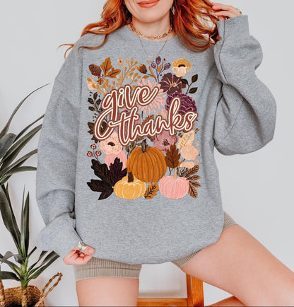 A woman wearing a grey sweatshirt with a graphic design of autumn leaves, pumpkins, and flowers, along with the text &quot;Give Thanks&quot; printed on it.