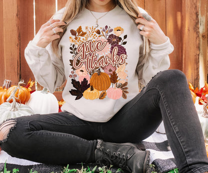 A woman wearing a white sweatshirt with a graphic design of autumn leaves and pumpkins, sitting on a blanket surrounded by pumpkins.