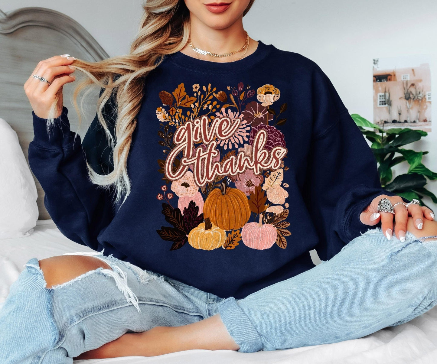 A woman wearing a navy blue sweatshirt with a colorful graphic design of pumpkins, flowers, and autumn leaves, sitting on a bed.