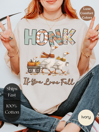 A woman wearing a white t-shirt with a graphic design of two geese and a pumpkin carriage, along with the text &quot;HONK If You Love Fall&quot;. She is making a peace sign with her fingers.