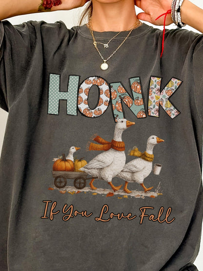 a person wearing a gray t-shirt with a graphic design depicting two white geese walking on a road with a pumpkin carriage, and the text &quot;HONK&quot; and &quot;If You Love Fall&quot; printed on the shirt.