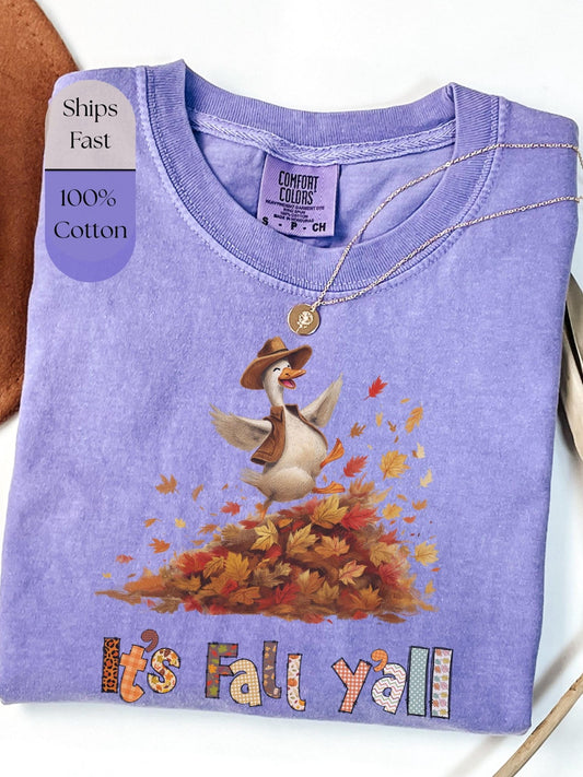 A purple t-shirt with a cartoon goose wearing a cowboy hat and surrounded by autumn leaves, with the text &quot;It&#39;s Fall y&#39;all&quot; printed on it.